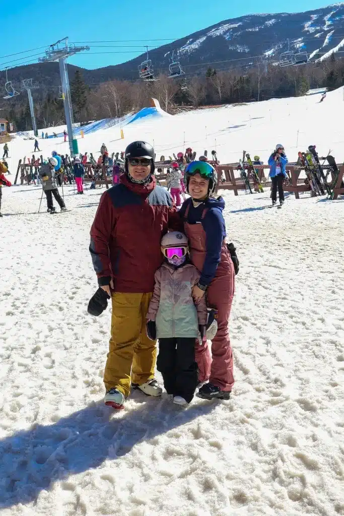 A family of three in ski gear poses together on a snowy ski slope with mountains, ski lifts, and other skiers in the background.