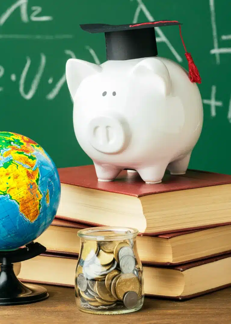 A piggy bank with a graduation cap sits on stacked books beside a globe and a jar of coins, in front of a green chalkboard.