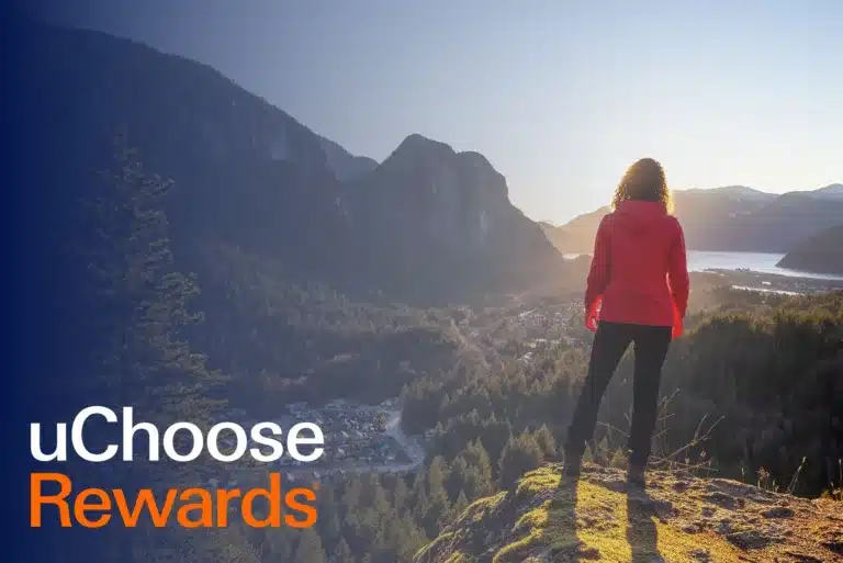 A person in a red jacket stands on a hilltop overlooking a scenic valley and mountains at sunrise; text reads uChoose Rewards.