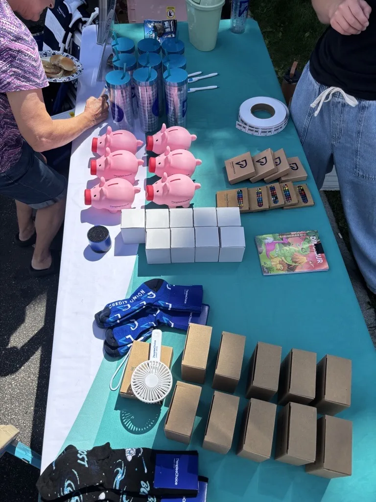 An overhead view of a teal table with piggy banks, boxes, socks, tumblers, pens, a fan, and brochures arranged in neat rows, with two people standing nearby.