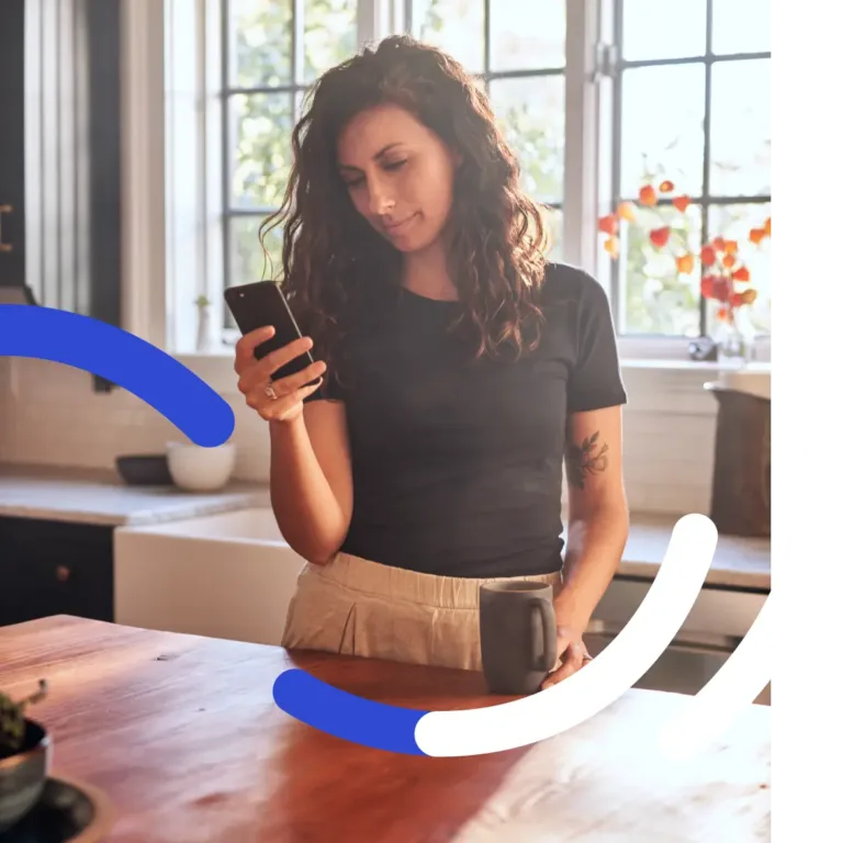 A woman holding a smartphone and a mug while standing in a kitchen.