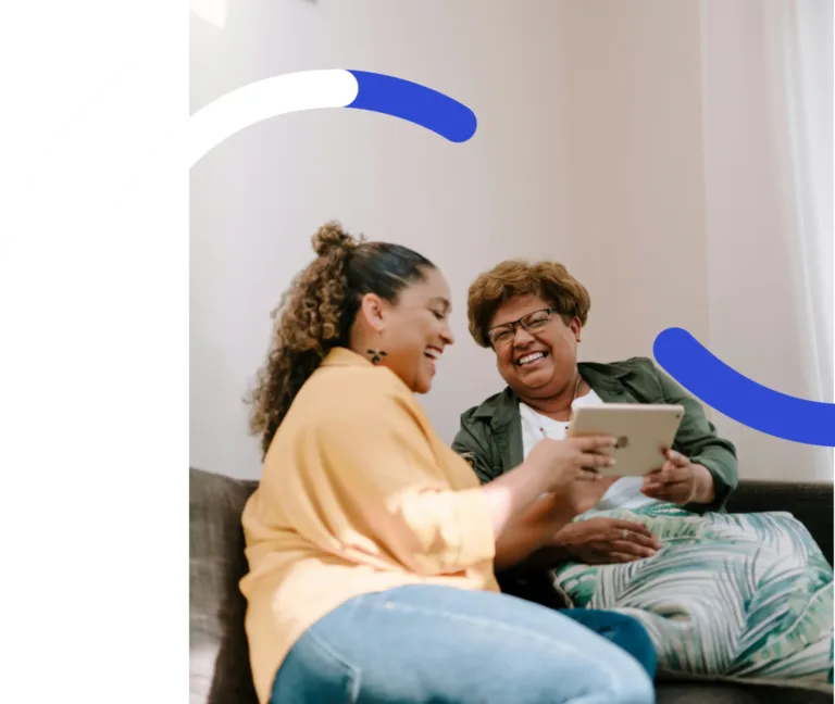 Two women sitting on a couch laughing while looking at a tablet.