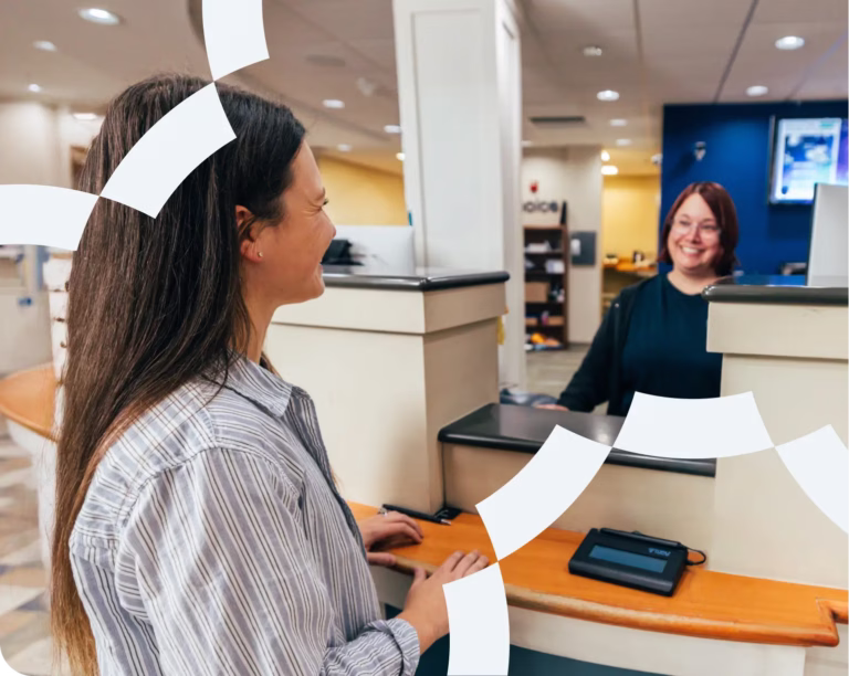 A customer stands at a bank or office counter, speaking with a smiling employee behind the desk.